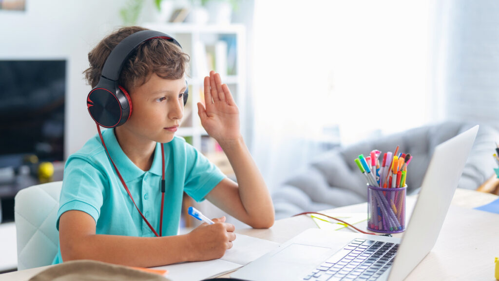 young student wearing headphones raises hand in front of laptop for online tutoring